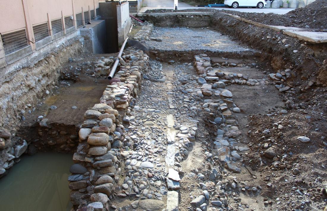 En primer terme, el carrer d’època baixmedieval amb les restes de les construccions que el flanquejaven a la dreta; al fons, la capa superior, la plaça o pati del segle XVII.