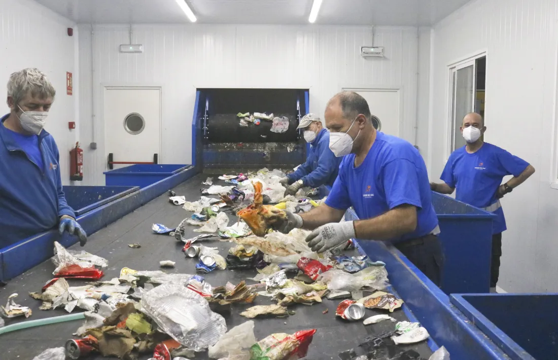 Treballadors de la planta de triatge d’envasos retiren impropis, un os de pernil, a la cinta de distribució.