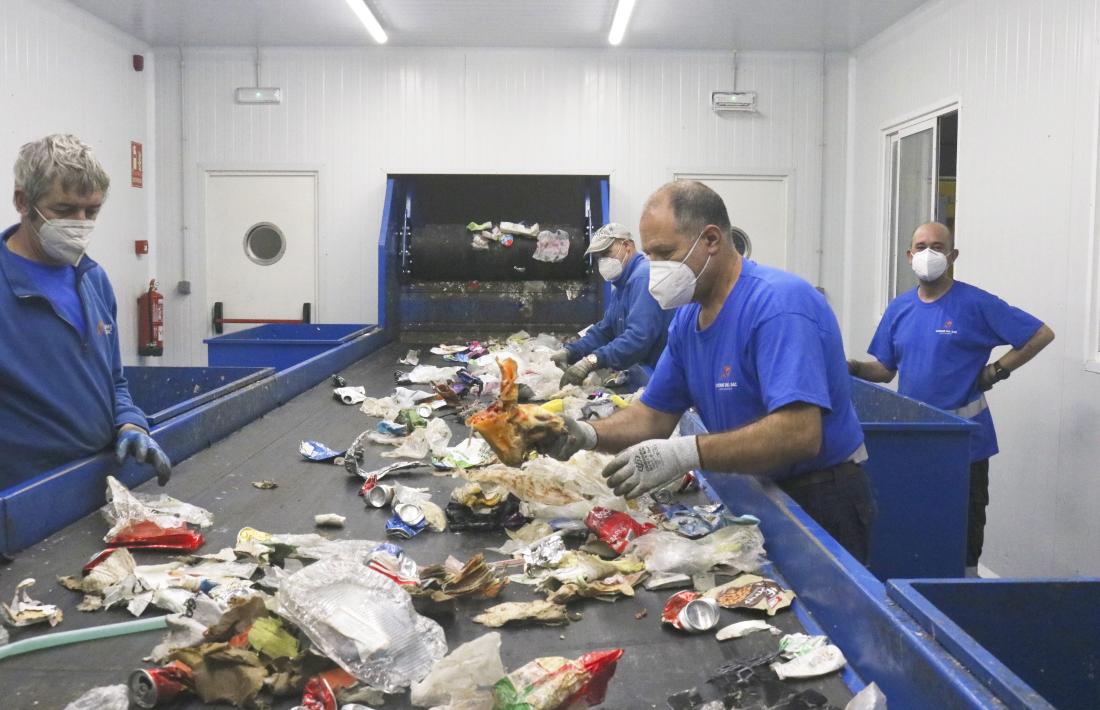 Treballadors de la planta de triatge d’envasos retiren impropis, un os de pernil, a la cinta de distribució.